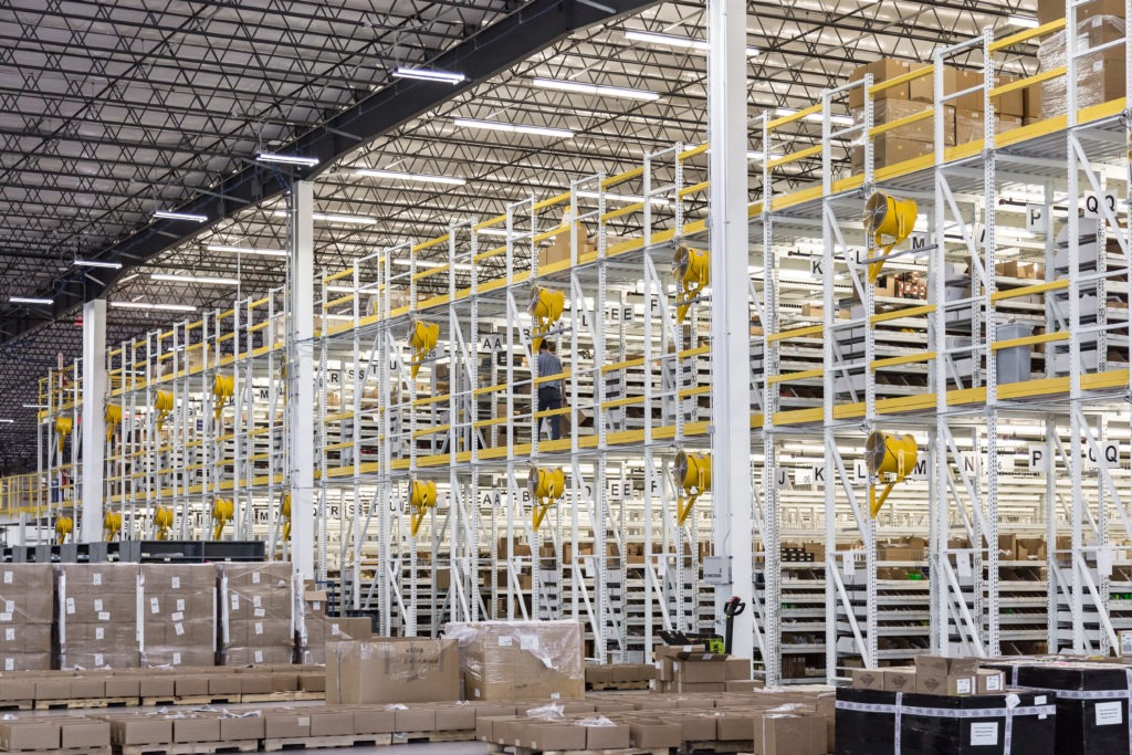 Interior of a large warehouse with multi-level white steel storage racks, yellow safety railings and equipment reels, stacked cardboard boxes on pallets, and a worker standing on an upper mezzanine level.